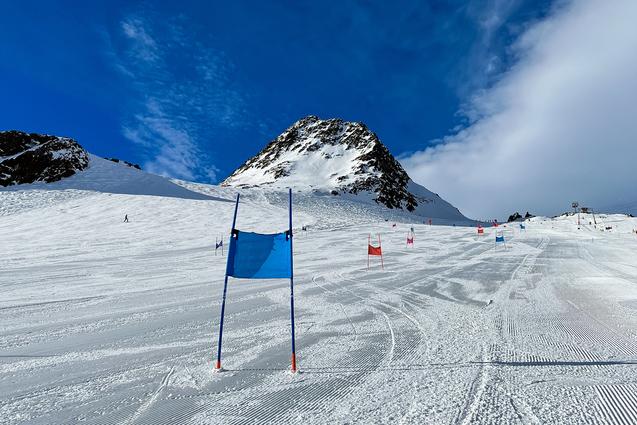 Skifahrer-Piste mit blauen und roten Toren, im Hintergrund ein schneebedeckter Berg unter blauem Himmel.
