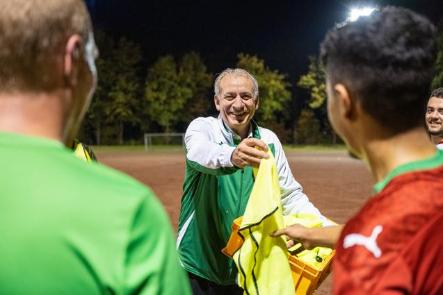 Trainer reicht gelbe Weste an Spieler während eines nächtlichen Fußballtrainings auf einem Platz.
