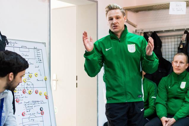 Trainer erklärt Taktik an Taktiktafel, Spieler hören im Hintergrund aufmerksam zu, in Umkleidekabine mit grüner Kleidung.